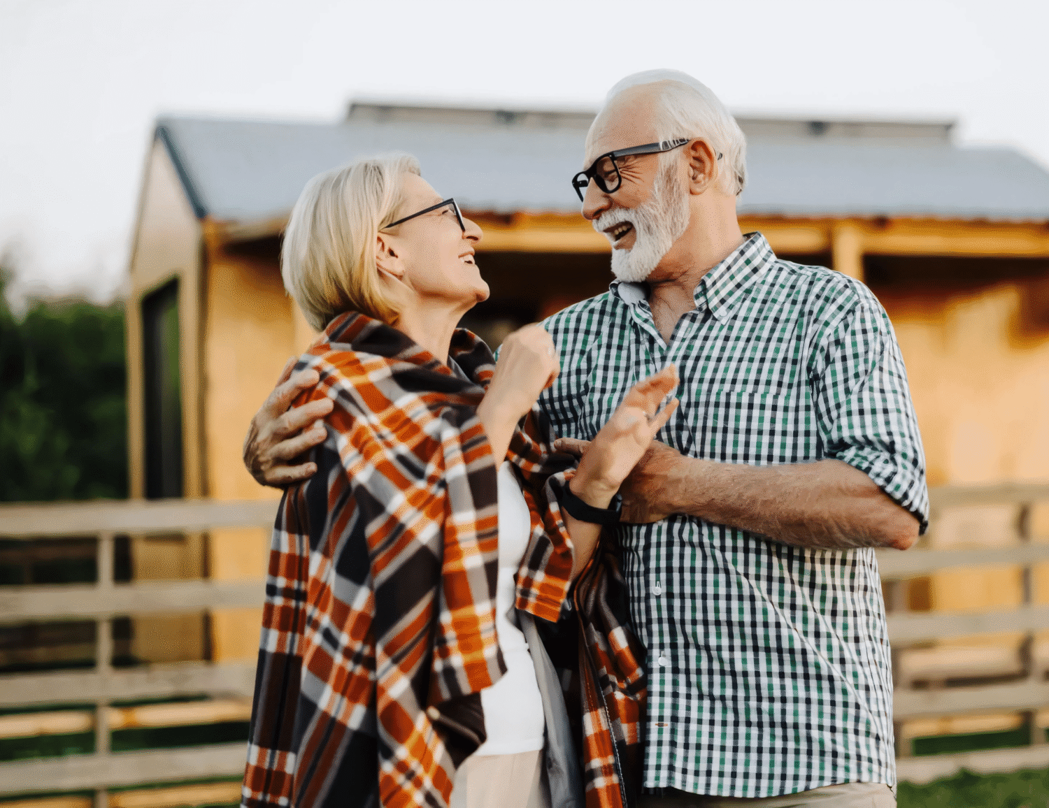 Couple laughing and embracing on a wooden porch, the person on the left wrapped in an orange plaid blanket and the person on the right wearing a green checkered shirt and glasses