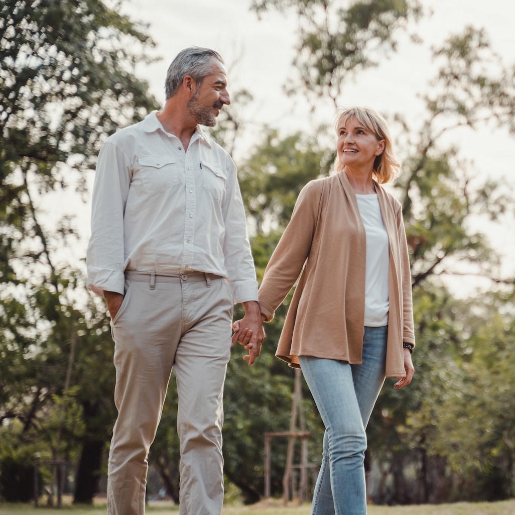 Man and woman holding hands while walking in a tree-lined park, both smiling and making eye contact.