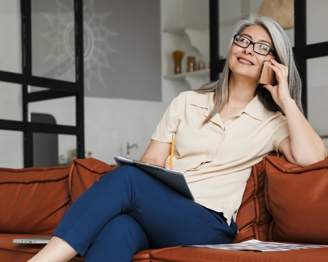 Gray-haired woman wearing glasses talks on a smartphone while writing on a clipboard on a sofa